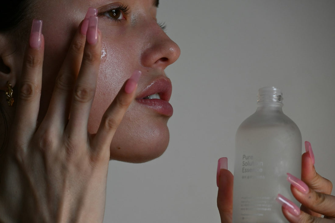 Mujer aplicando sérum en la mejilla junto a un frasco de vidrio